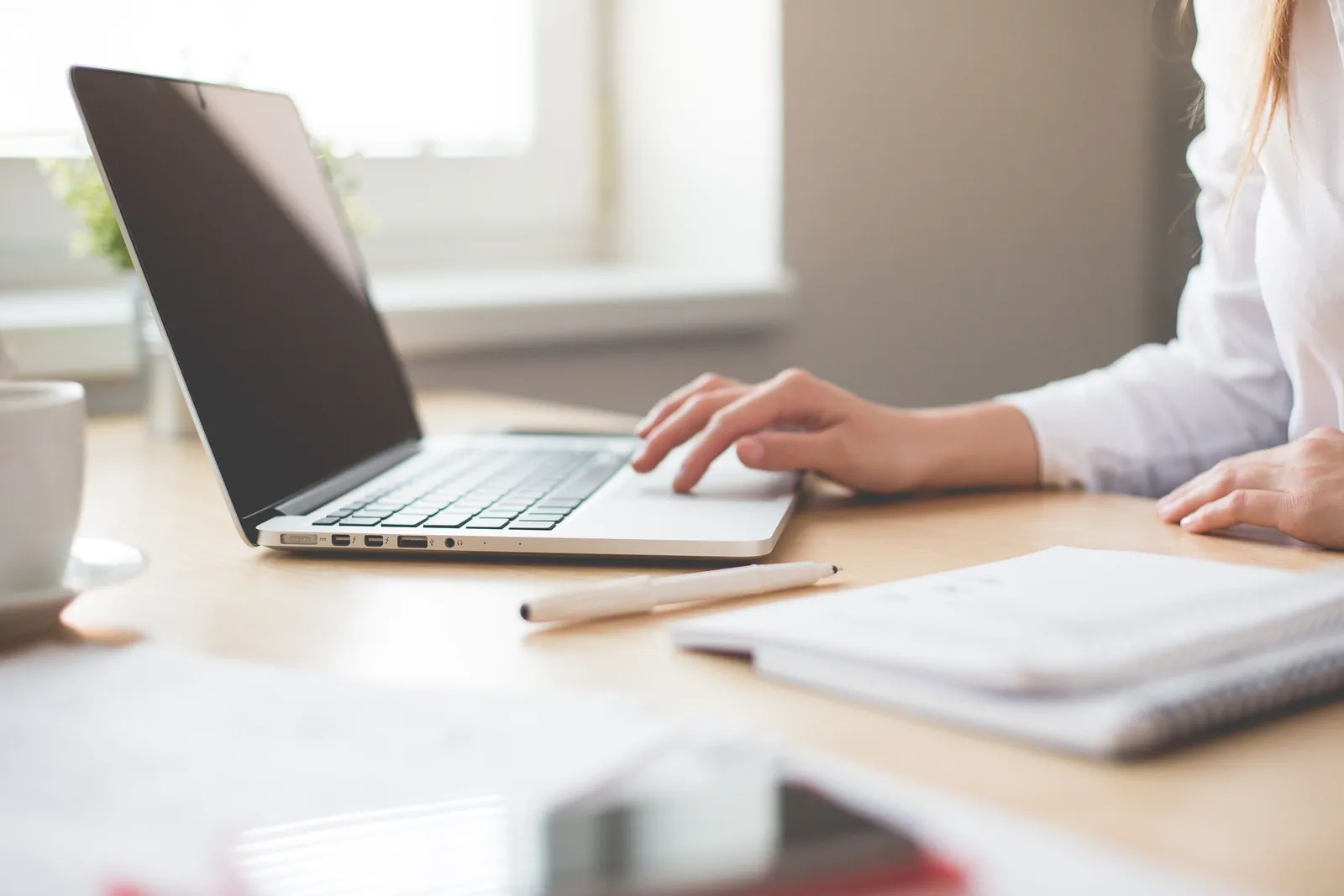 A businesswoman typing on a laptop at a bright indoor workspace with notebooks and a pen