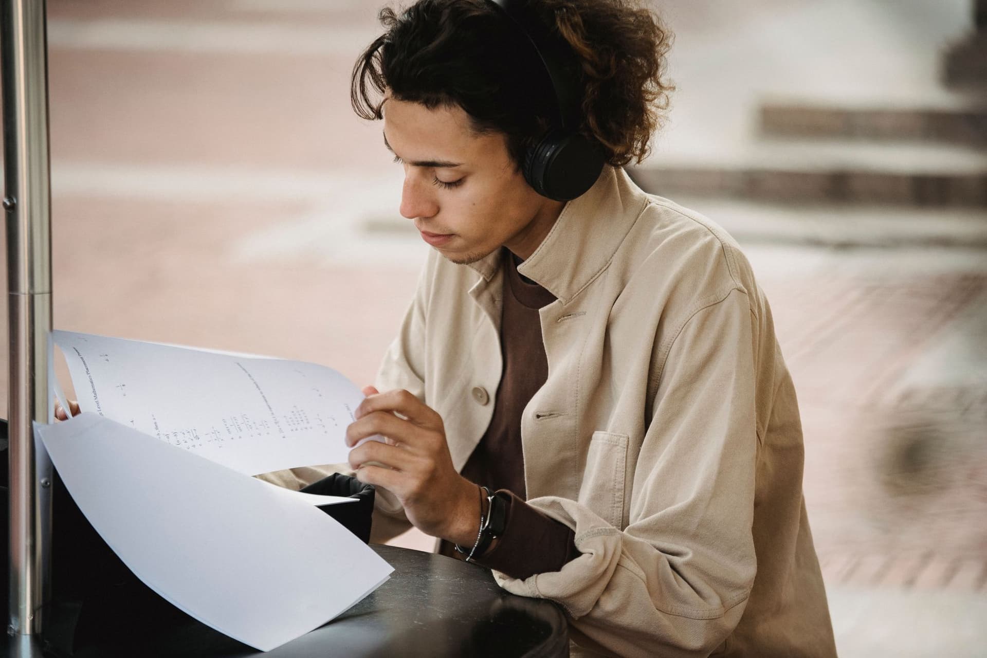 A young man wearing headphones reading a printed document at an outdoor table